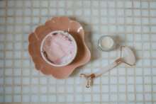 Close-up of spa essentials including pink cream, clay mask and jade roller on a tiled surface.
