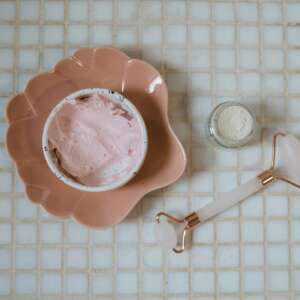 Close-up of spa essentials including pink cream, clay mask and jade roller on a tiled surface.